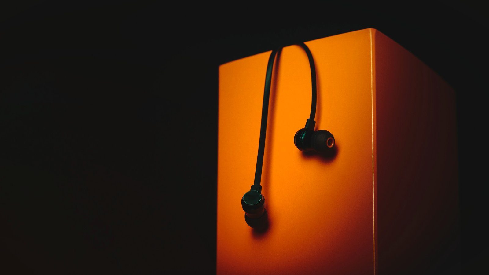 Unos auriculares de botón con cable colgados de una caja naranja, sobre un fondo oscuro